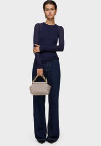 Kobieta ubrana w granatową bluzkę z długim rękawem i ciemne, szerokie jeansy, trzyma beżową pikowaną torebkę na łańcuszku w złotym kolorze.
