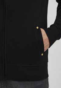 Schwarze Zip-Hoodie aus weichem Stoff, mit geripptem Saum und zwei seitlichen Taschen mit gelben Knöpfen. Nahaufnahme des Taschenbereichs.