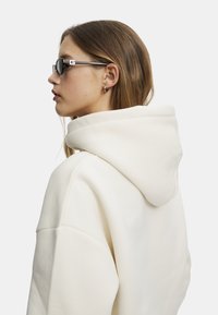 Krämfärgad oversized hoodie med stor huva och ribbad mudd, tillsammans med smala svarta solglasögon med detaljer i silver.