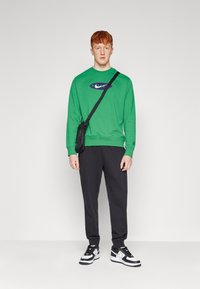 Sweatshirt verde de mangas longas com um logótipo, calças pretas e ténis em preto e branco. O modelo tem uma mala transversal e cabelo curto vermelho.
