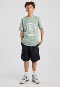 Mint green t-shirt with a white graphic text and broccoli illustration, paired with black cargo shorts and white sneakers with green accents.