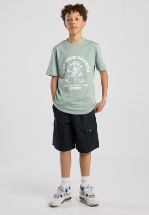 Mint green t-shirt with a white graphic text and broccoli illustration, paired with black cargo shorts and white sneakers with green accents.