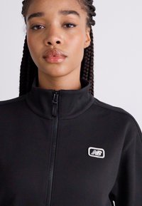 Schwarze Performance-Jacke mit Reißverschluss, hohem Kragen und einem weißen New Balance-Logo auf der linken Brust. Glatte Textur, sportlicher Schnitt.