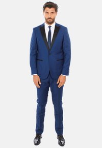 Abito blu navy con revers in satin nero, due tasche frontali e design strutturato; abbinato a una camicia bianca e una cravatta blu navy, completato da scarpe lucide.