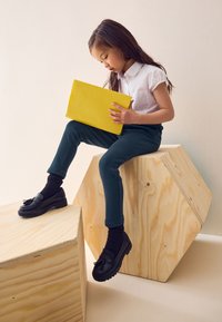 Bambino che indossa una maglietta bianca a maniche corte e pantaloni verde scuro, tiene in mano un tablet giallo. Scarpe nere con nappine; blocchi di legno usati come sedute.