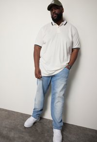 Camisa polo blanca con cuello y ribete de las mangas en contraste, combinada con jeans de mezclilla azul claro y zapatillas blancas. Complementado con una gorra verde.