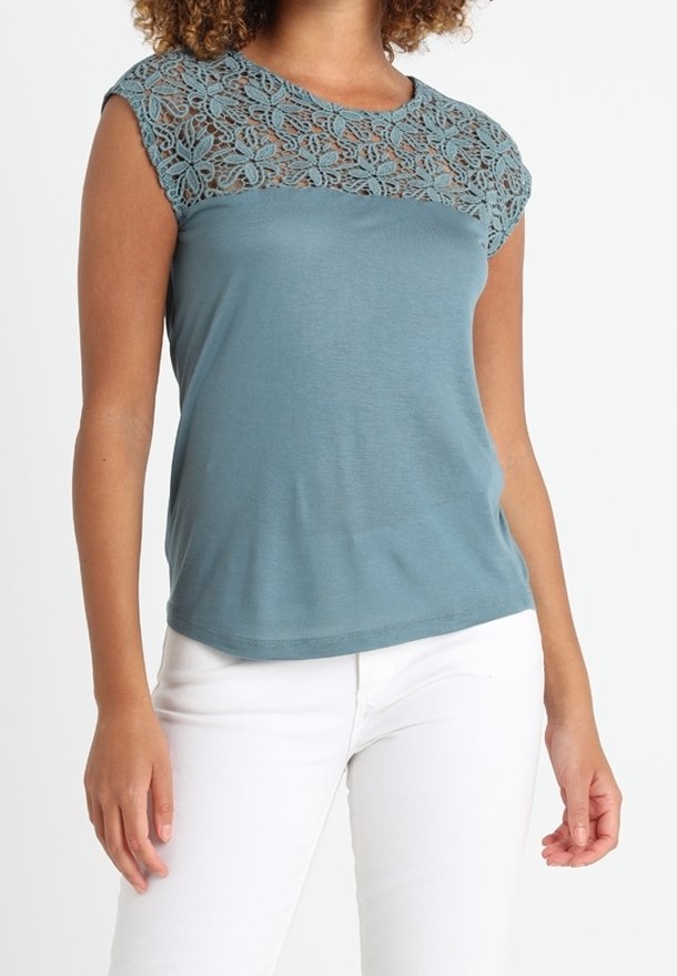 Femme portant un haut bleu canard à manches courtes avec un empiècement en dentelle florale, associé à un pantalon blanc, debout sur un fond blanc uni.