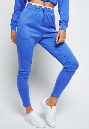 Person wearing bright blue joggers, white sneakers, and a blue cropped sweatshirt, standing against a plain background.