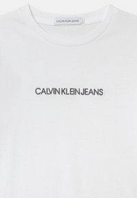 Valkoinen pyöreäkauluksinen t-paita, jossa on mustalla brodeerattu "CALVIN KLEIN JEANS" rinnan kohdalla ja brändilappu kauluksessa.