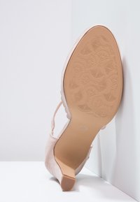 Beżowy zamszowy but na wysokim obcasie z kwiecistym wzorem na podeszwie i spiczastym czubkiem, ustawiony pionowo na białym tle w rogu.