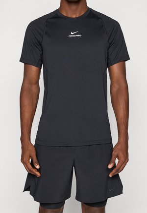 Mężczyzna w czarnej sportowej koszulce Nike Pro z krótkim rękawem i czarnych warstwowych spodenkach sportowych, stojący ze zrelaksowanymi ramionami po bokach.