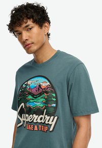 Camiseta de algodón en color teal que presenta un gráfico circular de montañas con colores vibrantes, y el texto "Superdry TAKE A TRIP" en letras negritas.