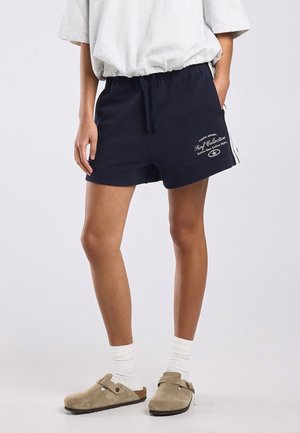 Persoon die marineblauwe korte broek met witte tekst, een lichtgrijs los T-shirt, witte sokken en beige instapschoenen draagt, staand op een lichte achtergrond.