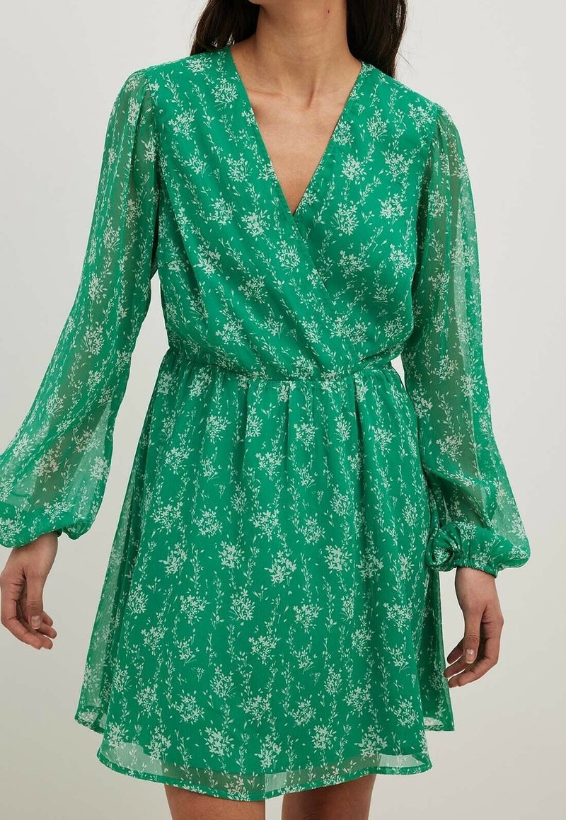 Robe portefeuille verte avec motif floral blanc, manches longues bouffantes transparentes, taille élastique et encolure en V portée par une personne.