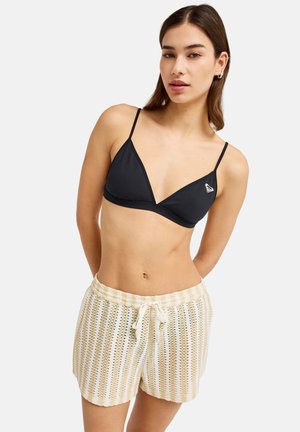 Giovane donna che indossa un top bikini nero a triangolo e pantaloncini di maglia a righe crema, posando con le braccia dietro la schiena su uno sfondo bianco neutro.