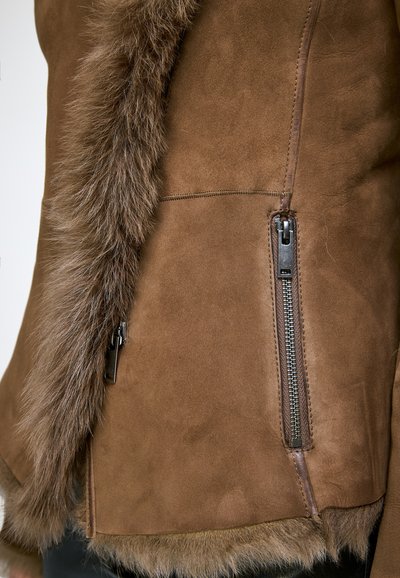 Veste en daim marron avec bordure en fourrure tout le long du bord et de l'ourlet, avec une poche avant zippée verticale en métal.