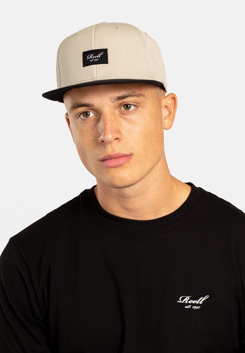 Gorra snapback de color beige claro con un parche cuadrado negro que presenta texto blanco. Llevada con una camiseta negra que muestra un bordado de logo blanco.
