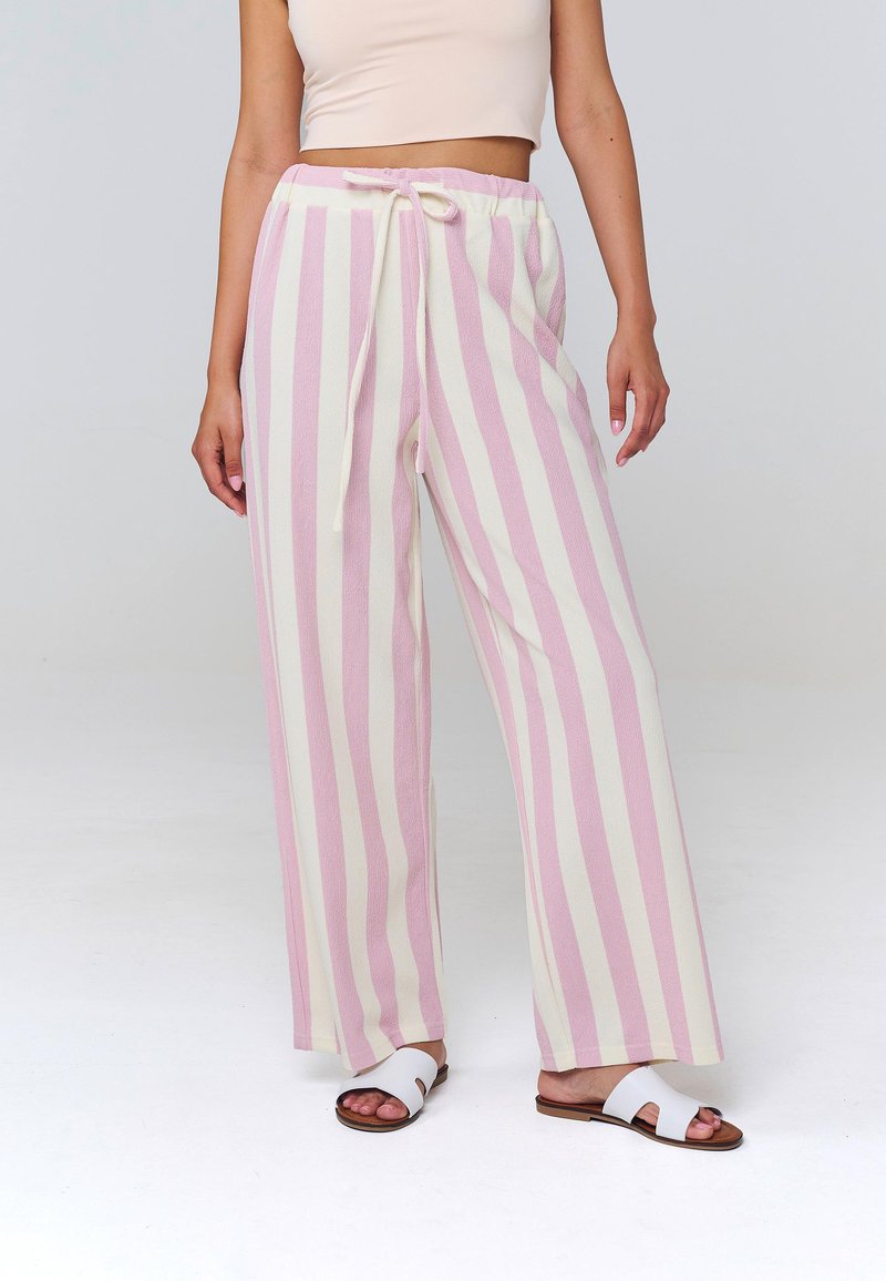 Femme portant un pantalon à jambes larges avec des rayures verticales roses et blanches, un top court blanc et des sandales blanches à enfiler.