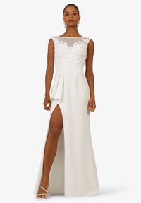 White evening gown with lace bodice and mesh neckline, featuring a high slit and draped, textured skirt. Simple strappy sandals.