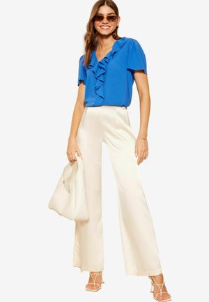 Top blu corto con dettagli a volant, abbinato a pantaloni bianchi a gamba larga. Accessoriato con una borsa color crema e tacchi beige con cinghie.