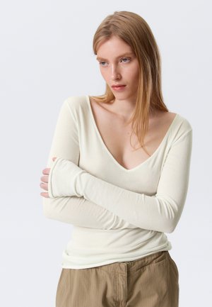 Noor naine pikkade blondide juustega, kandes kreemjat V-kaelusega pikka varrukaga pluusi ja beeže pükse, käed rüpes, vaadates kergelt alla ja küljele.