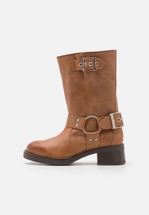 Hnědá kožená bota do poloviny lýtek se dvěma stříbrnými přezkami, detail ve tvaru kroužku na kotníku a nízkým blokovým podpatkem na bílém pozadí.