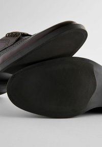 Chaussures en cuir marron avec un dessus lisse, des perforations décoratives et des semelles en caoutchouc noir dotées de motifs de traction texturés.