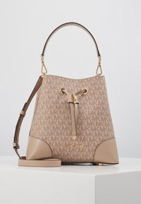 Sac seau beige Michael Kors avec un motif monogramme, matériel en ton doré, fermeture à cordon, poignée supérieure et bandoulière réglable sur une surface blanche.