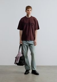 Brun oversize T-shirt med sort print, lyseblå jeans, sorte sko og en sort mulepose med en lilla sweater hængende over.