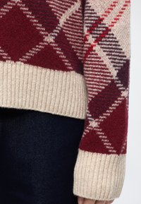 Manche de pull tricoté avec motif losanges rouge, blanc et violet foncé et bord-côte beige, portée sur un jean en denim foncé.