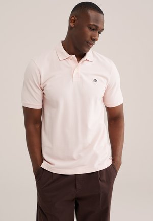 Muškarac u svijetlo roza polo majici s kratkim rukavima i malenim crnim logotipom na prsima te tamno smeđim hlačama, ruke u džepovima, gleda prema dolje u stranu.