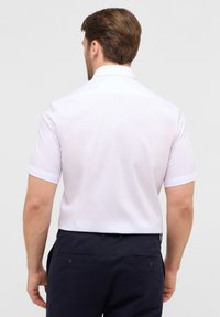 Witte short-sleeve shirt met een klassieke kraag en een gladde textuur; achteraanzicht met de schoudernaad en yoke zichtbaar. Gepaard met donkere broek.