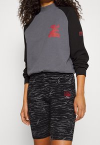 Grå och svart sweatshirt med hög krage och röd logotyp, kombinerad med svarta shorts som har ett grått abstrakt mönster och en logotext.