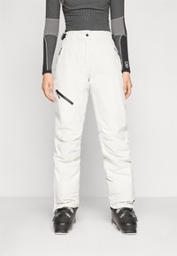 Hvite skibukser med avslappet passform, med glidelåslommer og forsterkede sømmer. Kombinert med svarte skistøvler. Teksturert stoff for isolasjon.