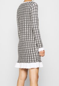 Abito lavorato a maglia in bianco e nero a scacchi, con maniche lunghe, orlo bianco volantes e tessuto strutturato. Cuciture visibili e silhouette aderente.