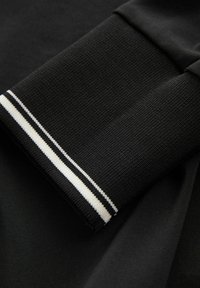 Poignet en tissu côtelé noir avec deux rayures blanches sur une manche de vêtement noire, montrant la texture et les détails du tricot.