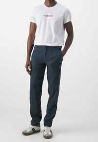 Hvid t-shirt med "PIZZICATO" i rød, navy blå linnedbukser og grå sneakers med hvide detaljer og et grønt element på sålen.