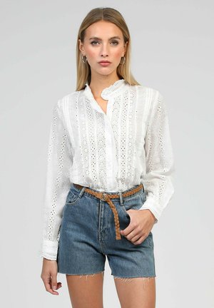 Femme portant un chemisier blanc en dentelle à manches longues, rentré dans un short en denim taille haute avec une ceinture tressée marron, debout devant un fond uni.