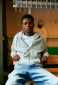 Grijze zipped hoodie over een witte shirt, gecombineerd met lichtblauwe jeans. Het model zit, handen in de zakken, voor een houten achtergrond.