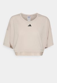 Jasnobeżowy krótki top wykonany z miękkiej tkaniny, z krótkimi rękawami i okrągłym dekoltem. Na piersi znajduje się czarne logo Adidas.