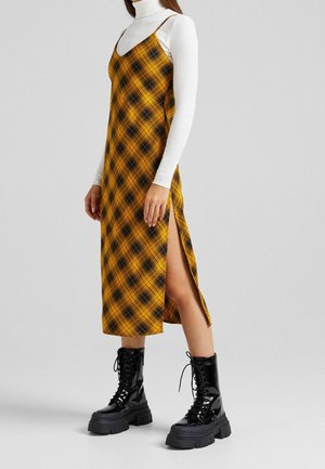 Mujer con botas negras de plataforma con cordones, suéter de cuello alto blanco de canalé y vestido amarillo de cuadros con abertura lateral.