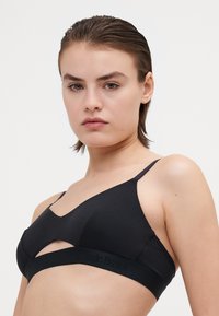 Soutien-gorge sans fil noir en tissu doux. Doté d'un décolleté en V et d'un design découpé. La bande élastique affiche le logo "Organic Basics".