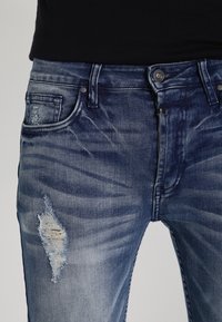 Niebieskie dżinsy z klasycznego denimu z wyblakłym wykończeniem, zniszczonymi detalami na udzie oraz klasycznym pięciokomorowym krojem z akcentami metalowymi.