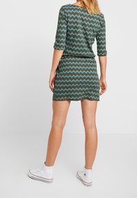 Zielona sukienka w zygzakowaty wzór w jasnym niebieskim kolorze, z rękawami trzyczwartkowymi i dopasowanym talią. W połączeniu z białymi trampkami za kostkę.