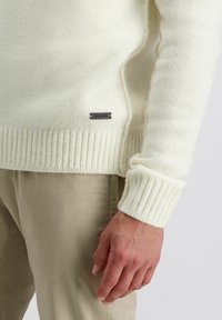Close-up van een persoon die een crèmekleurige gebreide trui draagt met geribbelde manchetten en onderrand, beige broek en een klein metalen merklabel op de trui.