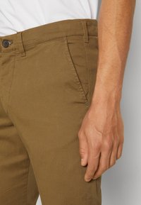 Pantaloni din bumbac maro cu o textură netedă, având o închidere cu nasture, buzunare pe laterale și o croială dreaptă. Mână vizibilă la talie.