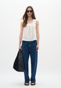 Haut blanc semi-transparent plissé avec des bretelles larges ; associé à un jean bleu droit taille haute. Le mannequin tient un grand sac fourre-tout noir.