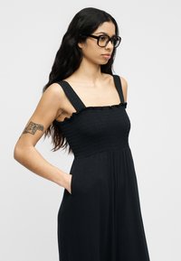Femme aux longs cheveux bruns et lunettes portant une robe noire sans manches avec un corsage froncé et des poches latérales, debout avec la main dans une poche.