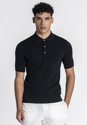 Młody mężczyzna z kręconymi włosami, ubrany w czarną krótki rękaw dzianinową koszulkę polo z drewnianymi guzikami i białe spodnie, stojący na jednolitym tle.