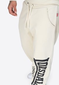 Beiga sweatpants gjorda av mjukt tyg, med dragsko i midjan, sidoficka och en stor svart logga tryckt längs benet.